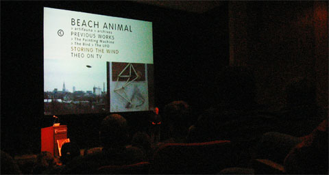 Theo Jansen on stage with screening in background and the public in the foreground